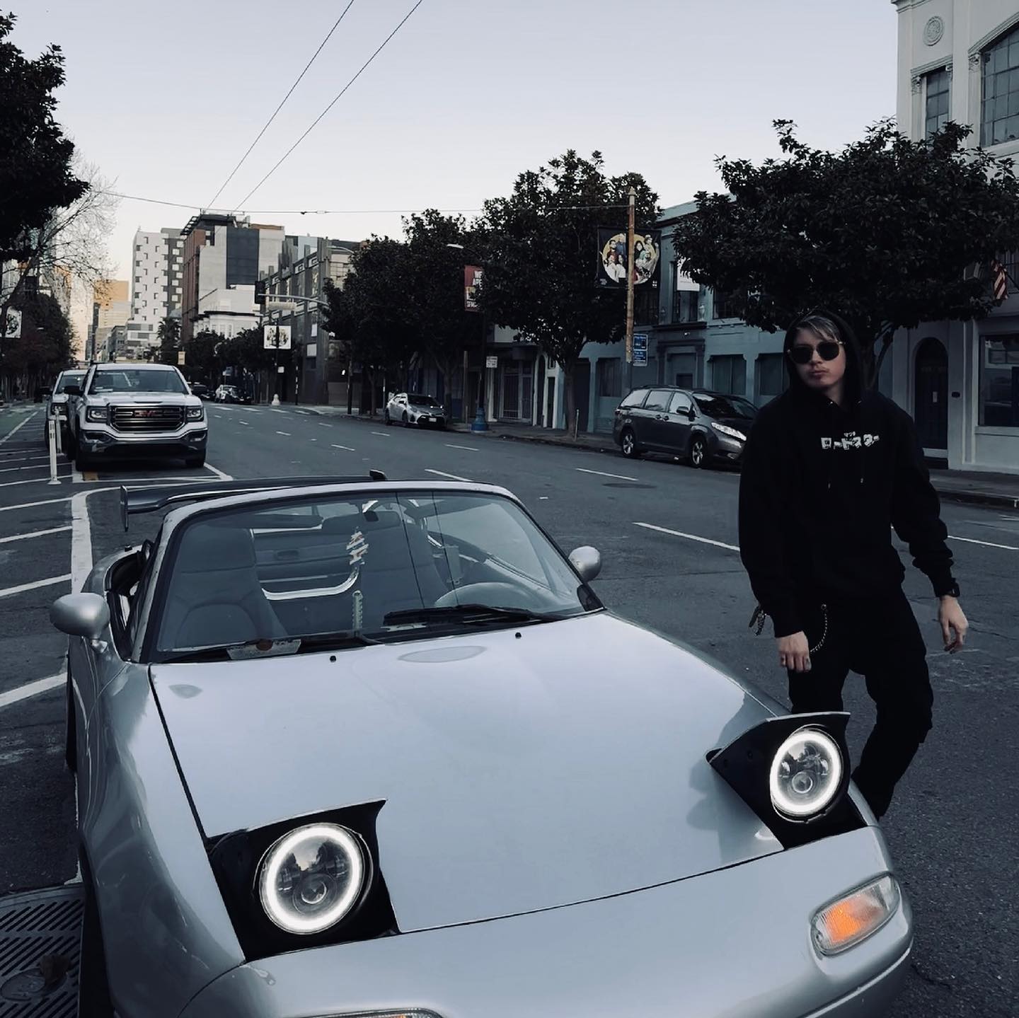 Person standing next to a light blue convertible car on a city street with a RevQor Hoodie.