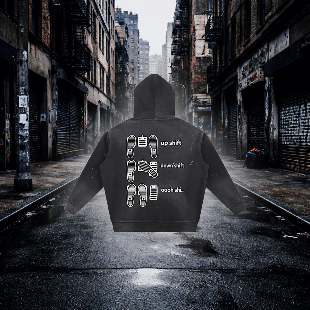 A black hoodie shown from the back, centered on a wet city street with closed storefronts and fire escapes in the background. The hoodie has white printed shift icons and the words 'up shift,' 'down shift,' and 'oooh shi.