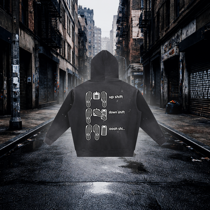A black hoodie shown from the back, centered on a wet city street with closed storefronts and fire escapes in the background. The hoodie has white printed shift icons and the words 'up shift,' 'down shift,' and 'oooh shi.