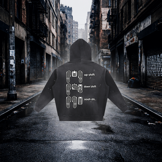 A black hoodie shown from the back, centered on a wet city street with closed storefronts and fire escapes in the background. The hoodie has white printed shift icons and the words 'up shift,' 'down shift,' and 'oooh shi.