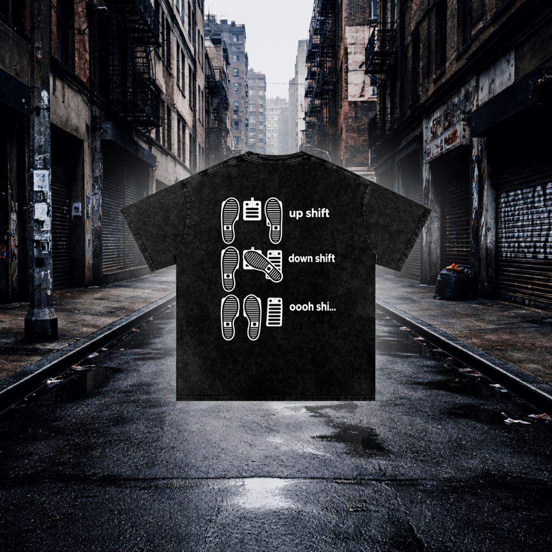 A black graphic t-shirt is superimposed over a moody, wet city street scene. The shirt features white line drawings of car footprints and gears with the text 'up shift', 'down shift', and 'ooh shi.