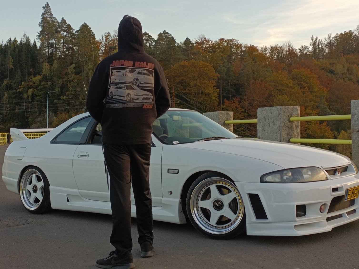 Person wearing a revqor hoodie with a car design, standing next to a white sports car in a forested area.
